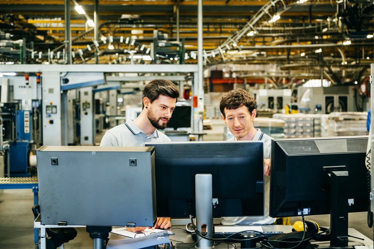 Two engineers standing behind several computer conitors in a huge factory and talking about problems of production line